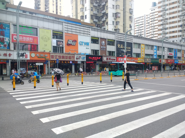 Eine belebte Stadtstraße mit Fußgängern an einer Ampelkreuzung, Gebäude mit Fenstern, Schilder, Laternen und Fahrzeuge, während einige Menschen Regenschirme in der Hand halten.