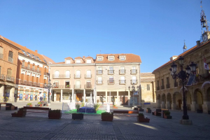 Ein Plaza Mayor in einem Stadtplatz mit einem zentralen Brunnen, umgeben von Bänken, Topfpflanzen, Straßenlaternen, einem Glockenturm und Gebäuden mit Fenstern unter einem klaren blauen Himmel.