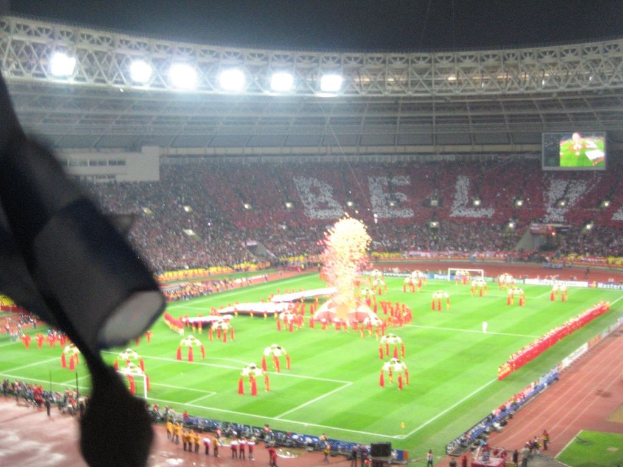 Ein Stadion mit Flutlicht, Eisengittern, Zuschauern, stehenden Personen, Werbetafeln, Anzeigetafeln, Überwachungskameras und dekorativen Fahnen.