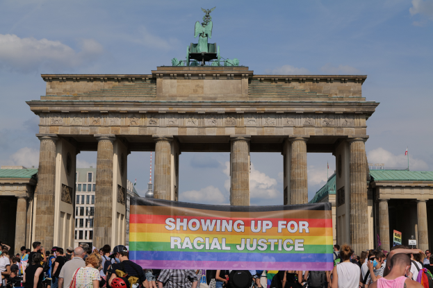 Eine Gruppe von Menschen steht vor dem Reichstagsgebäude in Berlin, Deutschland, und hält eine Fahne mit der Aufschrift "Rassengerechtigkeit". Das Gebäude hat Säulen und eine Statue, sowie Gebäude und einen bewölkten Himmel im Hintergrund.