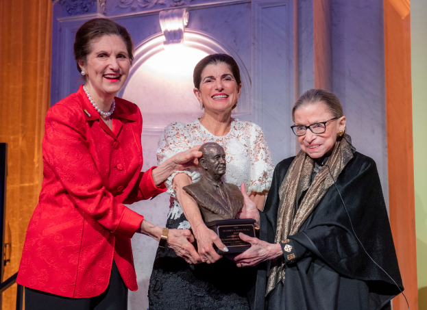 Drei Frauen stehen zusammen, wobei die mittlere Frau eine Statue hält, vor einer Wand mit einem Licht, einschließlich Ruth Bader Ginsburg und Nancy Pelosi, die mit einem Lebenswerk-Preis geehrt werden.