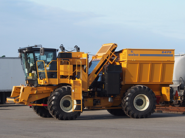 Großer gelber OXBO 2475 Radlader vor einem weißen Container auf einer Straße mit einem klaren blauen Himmel geparkt.