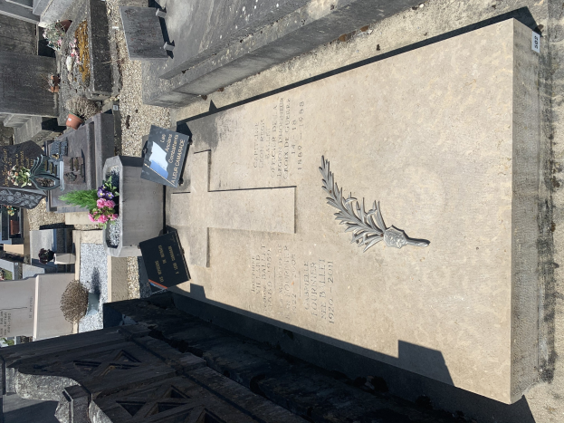 Ein Friedhof mit einem zentralen Grab, geschmückt mit einem Kränzchen, umgeben von Blumentöpfen und Pflanzen und einem Gebäude im Hintergrund.