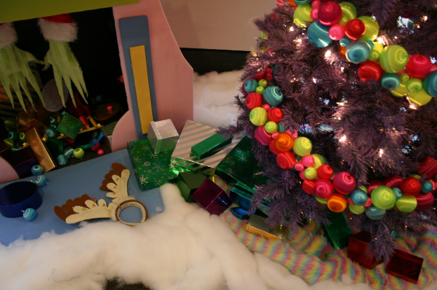 Drinnen steht ein geschmückter Weihnachtsbaum mit Lichterketten und Schmuck rechts, mit Geschenkboxen und Dekorationen in der Mitte, eine Hütte mit Dekorationen und Watte oben links und Watte und wollenes Tuch unten.