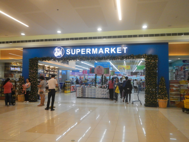 Eine Gruppe von Menschen, die vor einem Supermarkt stehen, einige halten Einkaufstaschen, mit einem Einkaufswagen, Topfpflanze, Gestellen mit Gegenständen, Schildern und einer Decke mit Deckenleuchten.