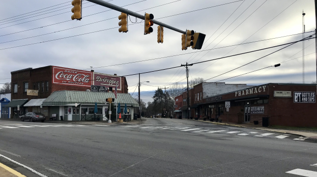 Eine Stadtstraße an einer Kreuzung mit einer Ampel, Fahrzeuge, Gebäude mit Beschilderung, Strommasten mit Kabeln, Bäume und ein bewölkter Himmel.