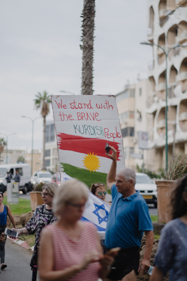 Eine Gruppe von Menschen marschiert nachts durch eine Straße, hält ein Schild mit der Aufschrift "Wir stehen zu den mutigen kurdischen Menschen" in der Hand, mit einem Van, der am Straßenrand geparkt ist, Straßenlaternen, Bäumen, Gebäuden und einem klaren blauen Himmel im Hintergrund.