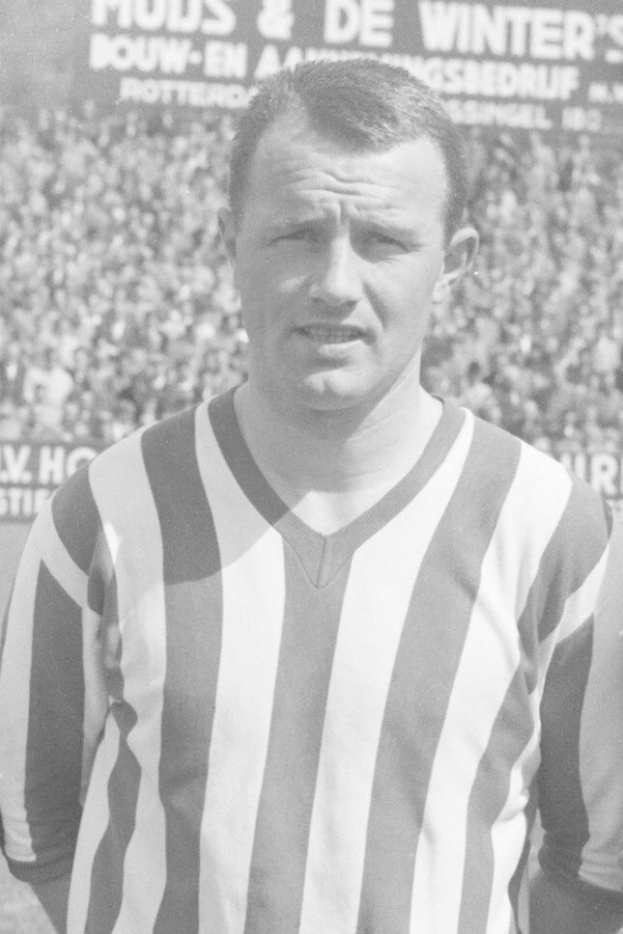 Ein Schwarz-Weiß-Foto eines Mannes in einem gestreiften Hemd vor einer Menge mit einer Tafel im Hintergrund, der legendäre niederländische Fussballspieler Bertrand.