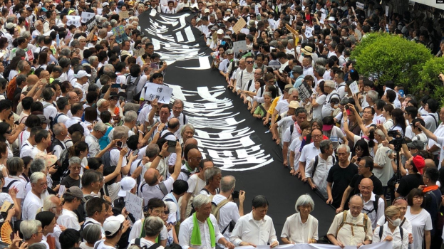 Eine große Gruppe von Menschen marschiert auf einer Straße, hält Protestschilder und Banner in der Hand, mit Grünzeug auf der rechten Seite und einem Gebäude im Hintergrund.