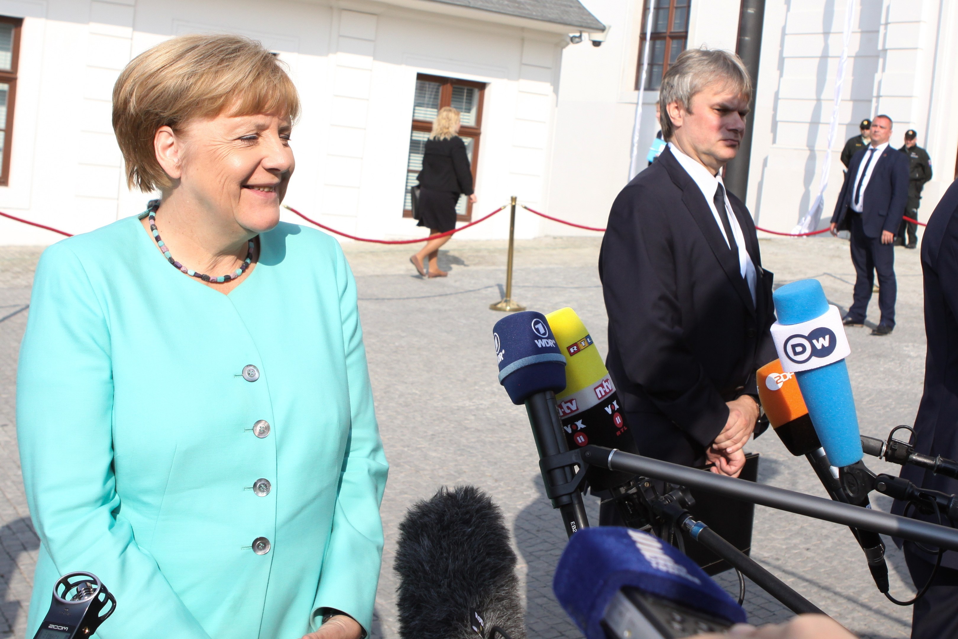Bundeskanzlerin Angela Merkel beim Sprechen mit Reportern vor einem Gebäude mit einer Gruppe von Menschen und Equipment, unter einem bewölkten Himmel.