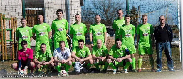Eine Gruppe von Männern in grünen T-Shirts steht auf einem Fussballfeld in der Nähe eines Torpfostens, mit einem Ball davor, während im Hintergrund Bäume, Gebäude und Fahrzeuge zu sehen sind.