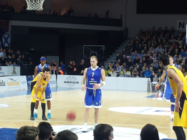 Eine Gruppe von Männern in blauen und gelben Uniformen, die Basketball auf einem Feld während eines 7days Eurocup Regular Season Round 2-Spiels zwischen Fenerbahce und Real Madrid spielen, mit Zuschauern auf den Tribünen und einem Basketballkorb am oberen Ende des Feldes.