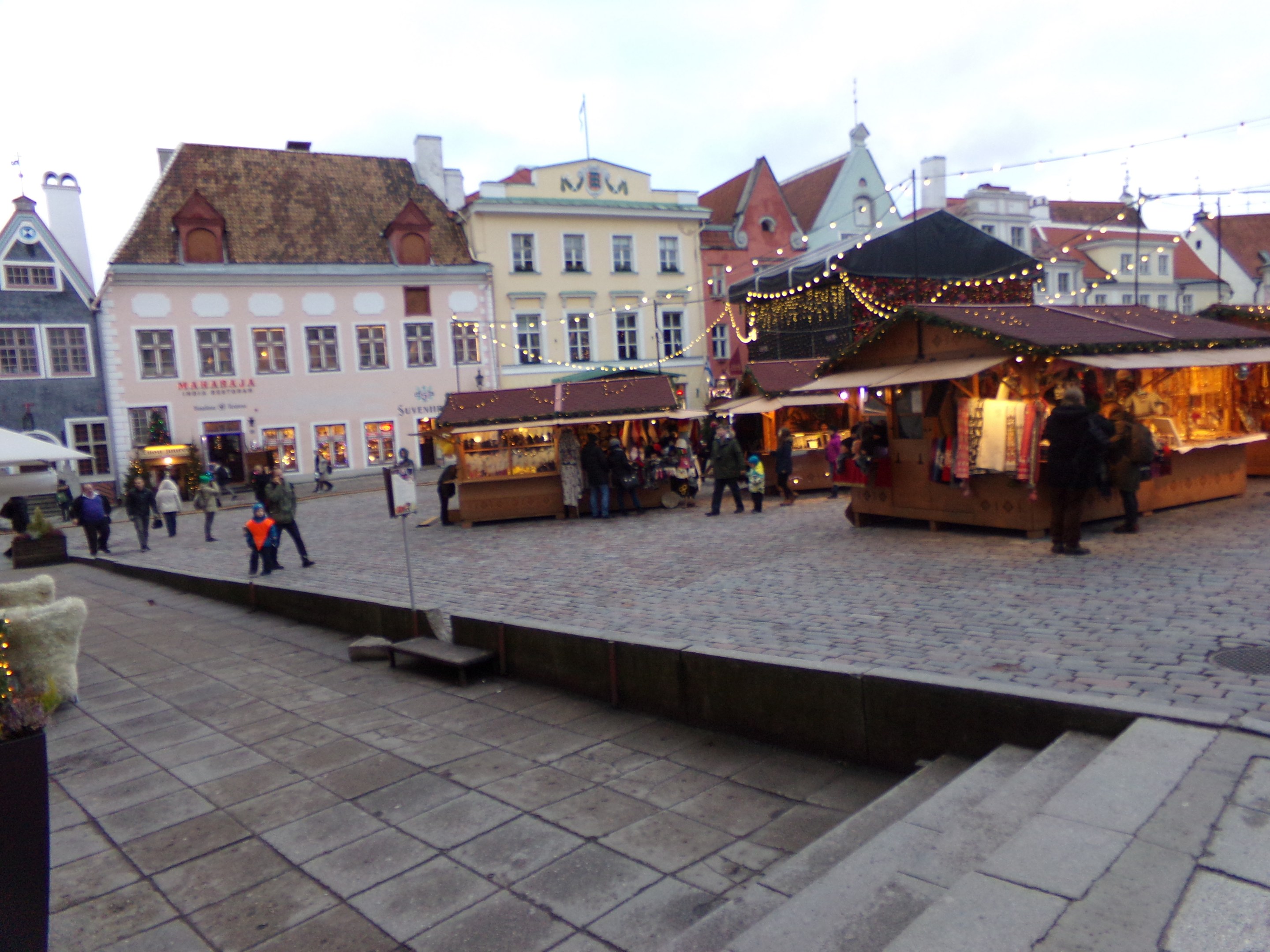 Ein geschäftiger Weihnachtsmarkt in Tallinn, Estland, mit Menschen um geschmückte Stände, festliche Lichter und Topfpflanzen, vor einem bewölkten Himmel und Gebäuden mit Fenstern.