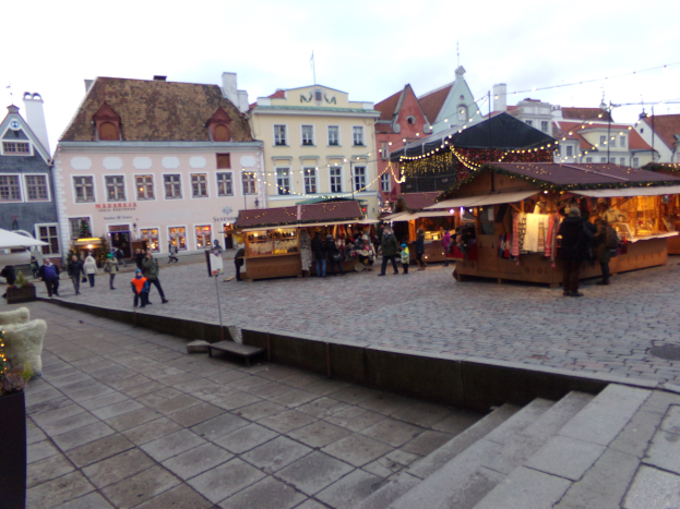 Ein geschäftiger Weihnachtsmarkt in Tallinn, Estland, mit Menschen um geschmückte Stände, festliche Lichter und Topfpflanzen, vor einem bewölkten Himmel und Gebäuden mit Fenstern.