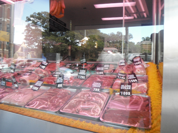 Eine Glasvitrine mit Tabletts voller Fleisch und Preisschildern, mit Gebäuden und Bäumen durch die Glasscheibe und einer Decke oben.
