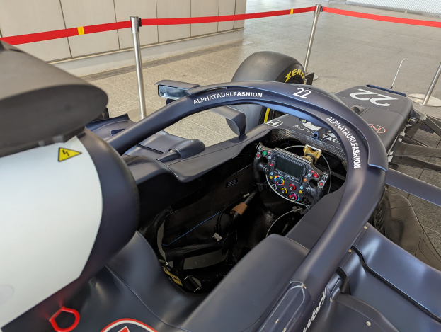Cockpit eines Formel-E-Autos in einer Ausstellung mit einem Lenkrad im Vordergrund, einer Wand im Hintergrund und um das Auto herum mit Bändern versehene Stangen.