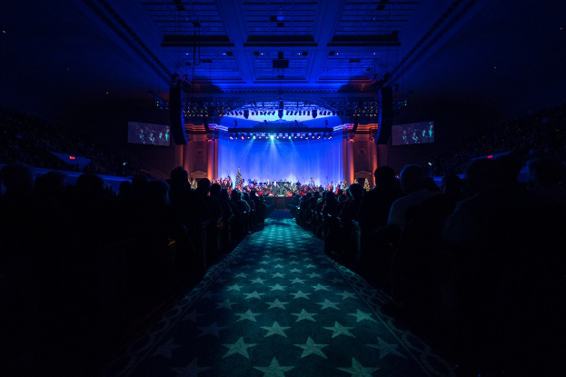 Ein großer Saal mit Menschen auf Stühlen, eine Bühne mit Musikern und eine Decke mit Lampen und Fernsehgeräten, was auf ein Konzerthaus während der Nationalkonvention 2019 hinweist.