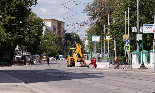 Eine Stadtstraße mit einer Baustelle in der Mitte, mit Fahrzeugen, Fußgängern, einem Radfahrer, Verkehrskegeln, Pfosten, Hinweisschildern, Strommasten mit Drähten, Bäumen, Gebäuden mit Fenstern und einem bewölkten Himmel im Hintergrund.