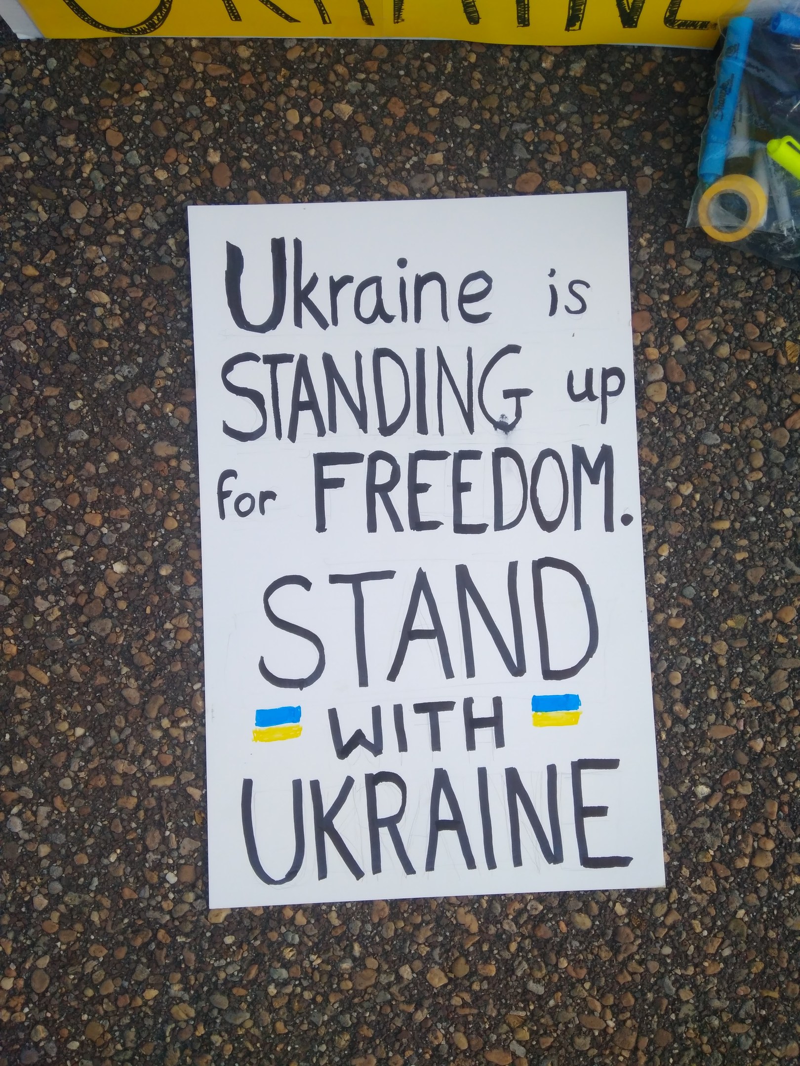 Ein weißes Papier mit dem Text "Ukraine steht für Freiheit ein, steht mit Ukraine" darauf, vor einem gelben Banner platziert, mit einer Plastikhülle mit verschiedenen Gegenständen rechts.