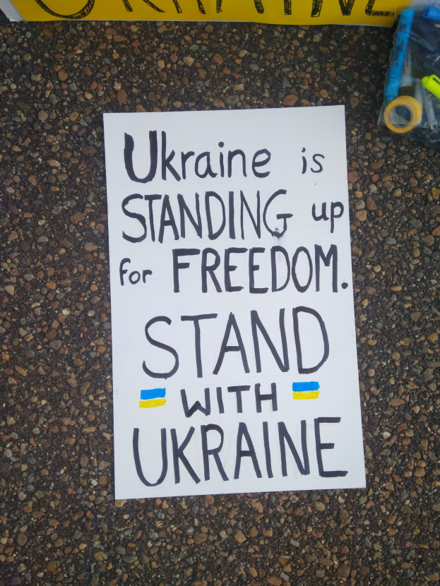 Ein weißes Papier mit dem Text "Ukraine steht für Freiheit ein, steht mit Ukraine" darauf, vor einem gelben Banner platziert, mit einer Plastikhülle mit verschiedenen Gegenständen rechts.
