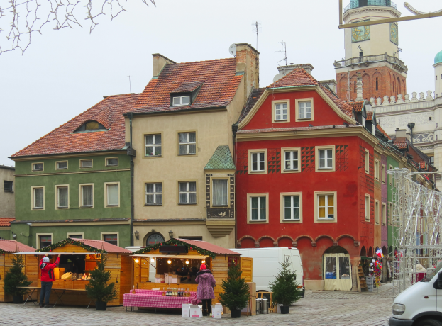 Ein lebendiger Weihnachtsmarkt im alten Stadtkern von Tallinn mit Menschen, Topfpflanzen, einem Tisch mit Gegenständen, einem geparkten Fahrzeug, Pfosten, einem Metallrahmen, Gebäuden mit Fenstern, einem Kirchturm, Antennen auf einem Dach und einem bewölkten Himmel.