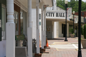 Ein Rathaus umgeben von Gebäuden, Straßenmasten, Straßenlaternen, Namensschildern, Kraftfahrzeugen, Absperrgittern, Stühlen, Topfpflanzen, Bäumen und einem bewölkten Himmel.