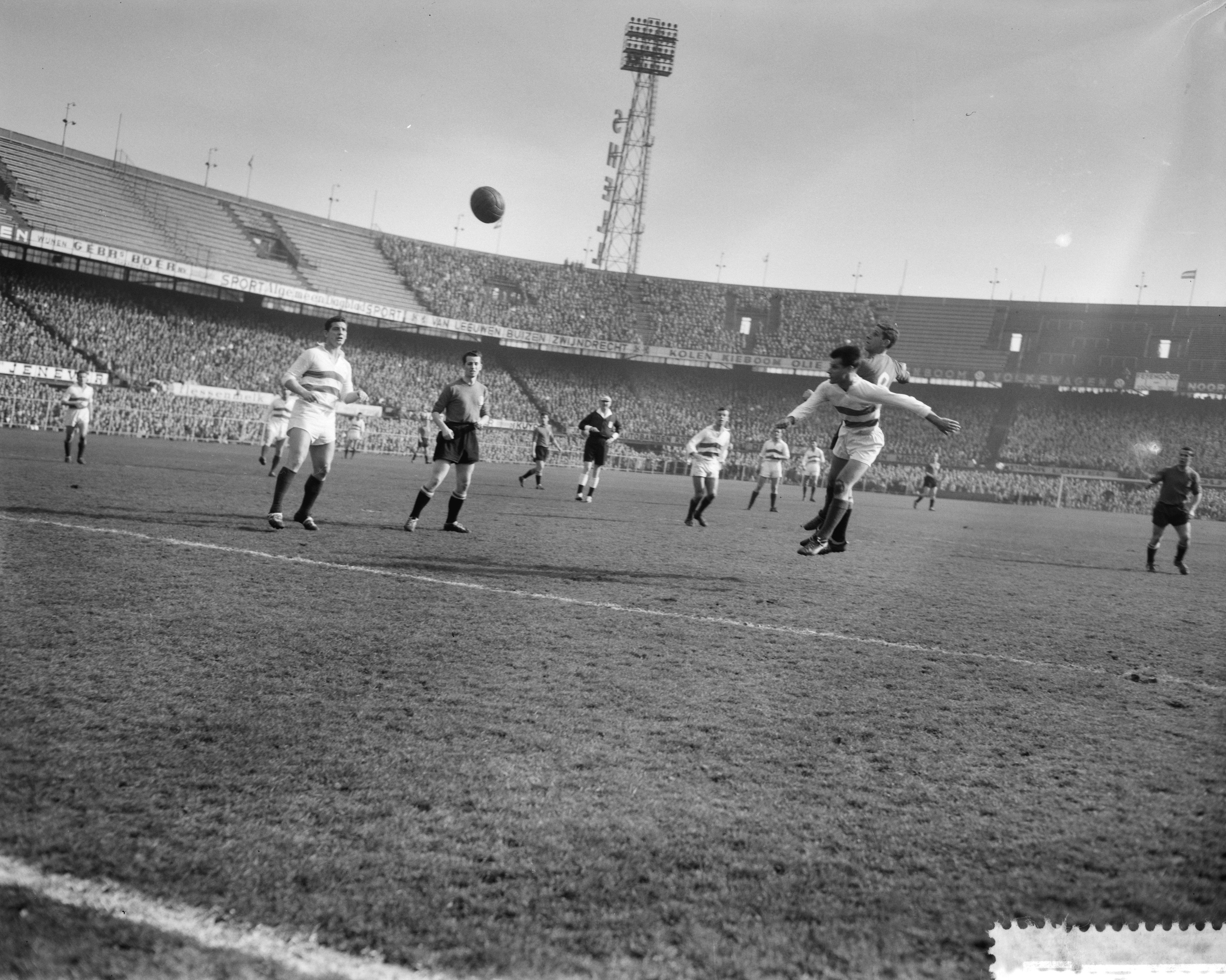 Eine Gruppe von Männern spielt Fußball auf einem Feld mit einem Stadion, Flutlicht und einem klaren Himmel im Hintergrund.