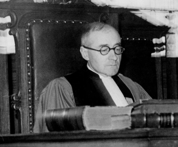 Ein Schwarz-Weiß-Foto eines Mannes mit Brille und Robe, der an einem Schreibtisch mit einem Buch sitzt und ernst schaut.