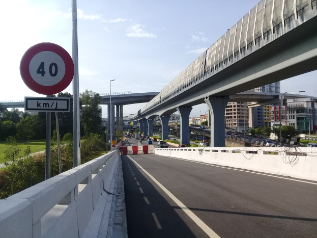 Autobahn mit Tempolimit-Schild, Fahrzeugen, Brückenpfeilern, Laternen, Bäumen, Gebäuden und bewölktem Himmel im Hintergrund.
