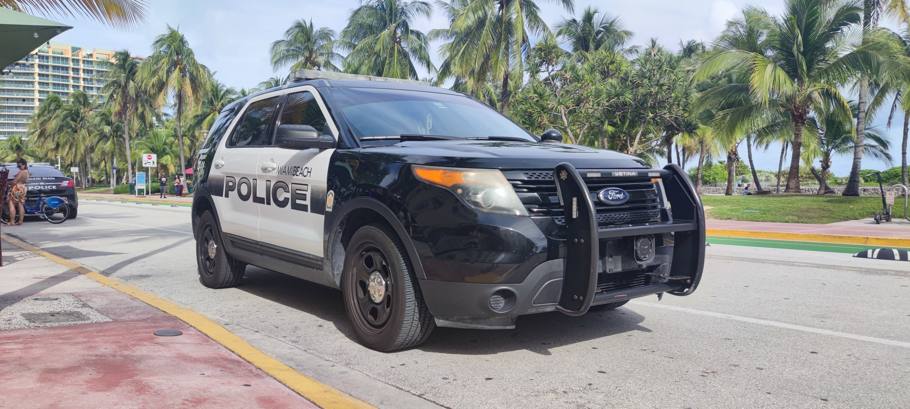 Polizeifahrzeug am Straßenrand geparkt mit Menschen, Fahrrädern, Gras, Bäumen, Gebäuden und einem klaren blauen Himmel im Hintergrund; Vorderrad zeigt Einsatzbereitschaft an.