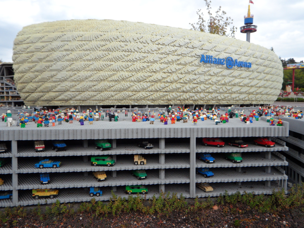 Großes Lego-Modell des Allianzstadions in München, Deutschland, mit Menschen davor, umgeben von Bäumen, Pfählen, einem Turm und Grün unter einem bewölkten Himmel.
