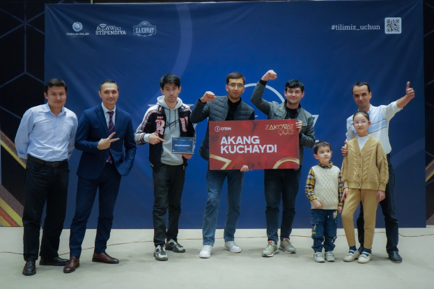 Gruppe von Menschen auf einer Bühne mit einer Tafel, auf der "Akang Kuchaydi - Zakovat Quiz" steht, im Hintergrund ein Banner, ein Barcode und Lichter