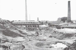 Ein Schwarz-Weiß-Foto einer Baustelle mit zwei Schornsteinen im Hintergrund, umgeben von Gebäuden, Masten, Bäumen und einem klaren Himmel, mit Text am unteren Rand, der darauf hinweist, dass es sich um ein kohlebetriebenes Kraftwerk handelt.