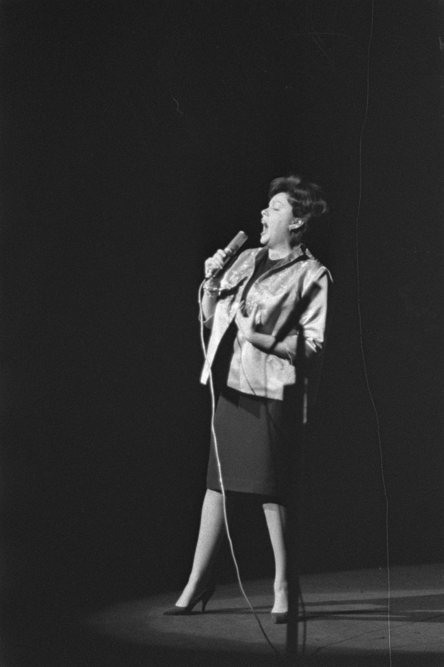 Eine Frau in einem schwarzen und weißen Outfit aus den 1950er-Jahren singt fröhlich auf einer von Scheinwerfern beleuchteten Bühne und hält ein Mikrofon.