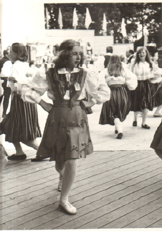 Eine Gruppe von Frauen in traditioneller bayrischer Tracht tanzt auf einem Holzboden, mit einer Wand, B├Ąumen und anderen Gegenst├Ąnden im Hintergrund. Text ist unten auf dem Bild sichtbar.