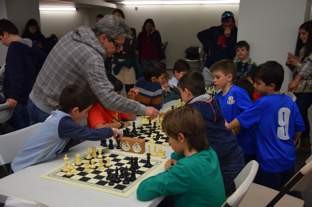 Kinder spielen Schach bei einem Wettbewerb, während Erwachsene von hinten und seitlich zuschauen.
