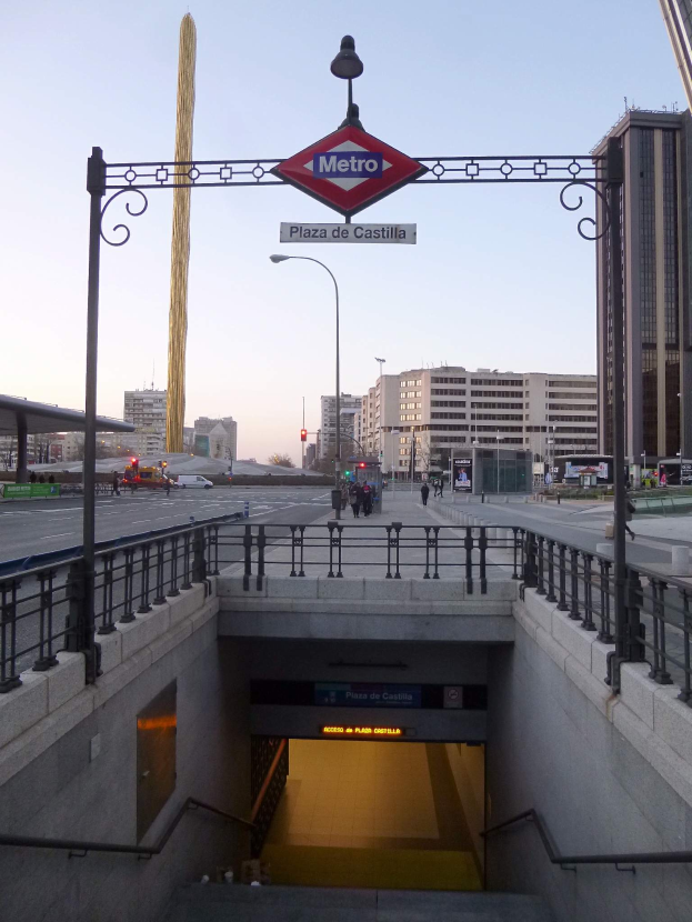 Eine belebte Stadtkulisse mit Gebäuden, Fahrzeugen, Fußgängern, einem U-Bahn-Eingang und einem Schild darüber bei klarem Himmel.