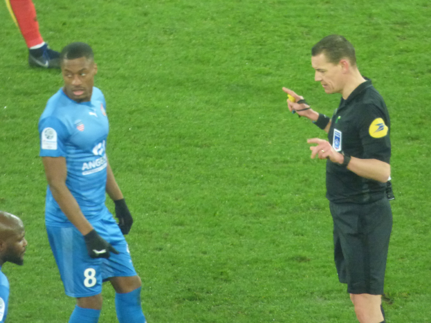 Schiedsrichter spricht mit einem Fußballspieler auf dem Feld, umgeben von einer Gruppe von Menschen, wobei eine Person ein Objekt in der Hand hält und die Beine einer anderen Person im Hintergrund zu sehen sind.