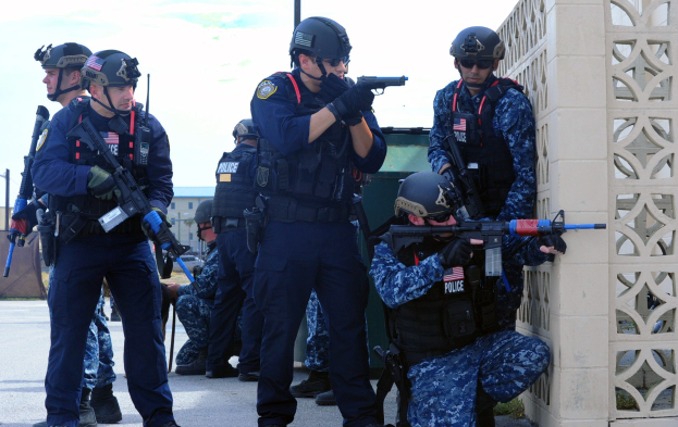 Gruppe von Polizisten in Uniform auf einer Brücke stehend, jeder trägt einen Helm und hält eine Waffe, mit einer Wand rechts und Gebäuden, Pfählen, Bäumen und Himmel im Hintergrund.