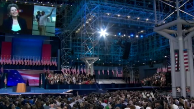 Eine große Menschenmenge sitzt vor einer Bühne mit einem Podium, einem Mikrofon, Flaggen, einem Bildschirm und Lichtausrüstung und wartet auf eine Debatte.