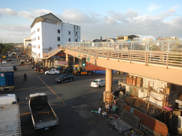 Eine belebte Stadtstraße mit zahlreichen Autos, Lastwagen und Fußgängern, eine Brücke mit Geländern und Säulen, Verkehrsampeln, Gebäude mit Fenstern, Bäume und ein bewölkter Himmel.