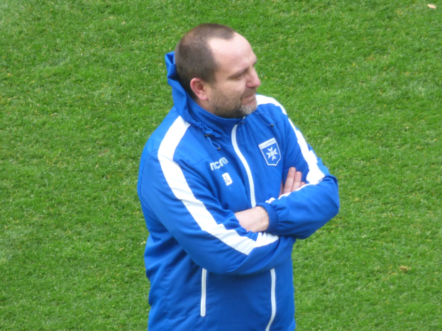 Ein Mann in einer blauen Jacke steht selbstbewusst auf einem grünen Feld und erscheint als Trainer.