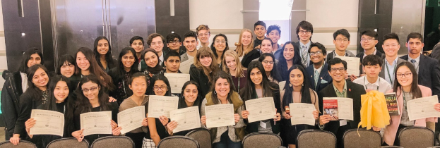 Gruppe von Menschen mit Urkunden, die in einem Raum mit Stühlen und einer Wand im Hintergrund posieren, wahrscheinlich bei einer Abschlussfeier für das akademische Jahr 2019-2020.