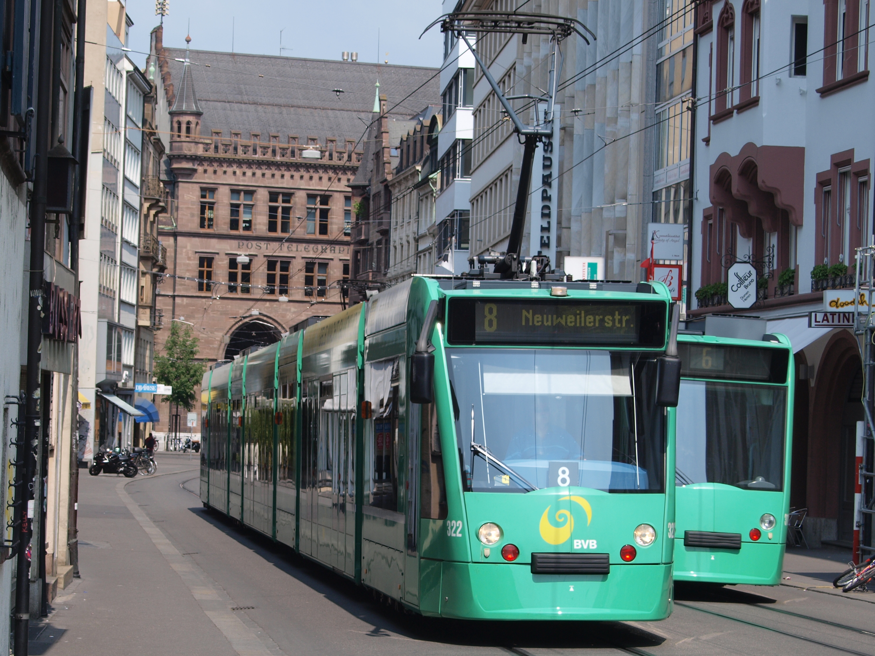 Zwei grüne Straßenbahnen fahren auf einer von Hochhäusern gesäumten Stadtstraße, mit parkenden Fahrrädern und Fußgängern auf den Gehwegen, einem Baum und einem klaren blauen Himmel im Hintergrund.
