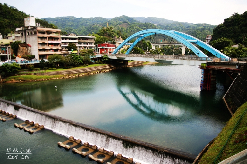 Eine Brücke überspannt einen Fluss mit Gebäuden, Bäumen und Hügeln im Hintergrund, unter einem klaren Himmel.
