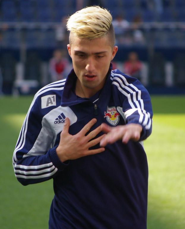 Ein junger Mann in einem blauen T-Shirt steht auf einem Fußballfeld und streckt die Arme aus, entschlossen wie ein Flügelspieler.