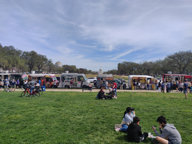 Eine Gruppe von Menschen auf einem grünen Feld beim Washington D.C. Food Truck Festival, umgeben von Fahrzeugen, Fahrrädern und anderen Gegenständen, mit Bäumen, Gebäuden und einem bewölkten Himmel im Hintergrund.
