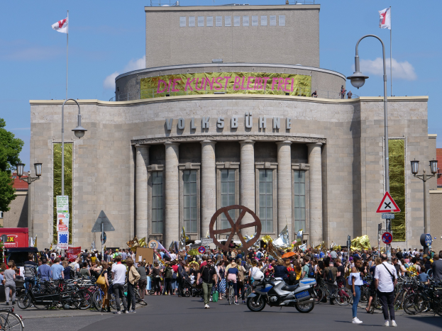 Eine große Gruppe von Menschen versammelte sich vor einem Gebäude mit Säulen und Text darauf, umgeben von Straßenmasten, Straßenlaternen, Hinweisschildern, Motorrädern, Kraftfahrzeugen, Bäumen und einem bewölktem Himmel, während einer Demonstration in Berlin, Deutschland.