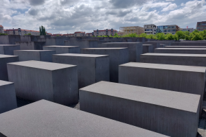 Betonschollen in einem Gittermuster am Holocaust-Mahnmal für die ermordeten Juden Europas in Berlin, mit Bäumen und Gebäuden im Hintergrund unter einem bewölkten Himmel.