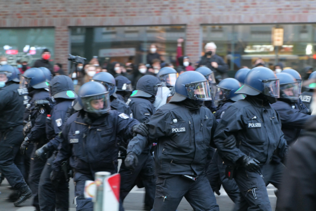 Eine Gruppe von Polizisten in Einsatzausrüstung, einschließlich Helmen und Handschuhen, die eine Straße entlanggehen mit einem unscharfen Hintergrund, der ein Gebäude mit Glaswänden und ein Schild an einem Pfahl zeigt.